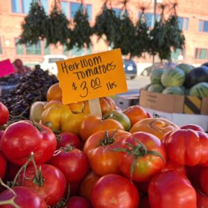 A pile of tomatoes with a sign that says Heirloom Tomatoes $3/lb and hanging herbs in the background